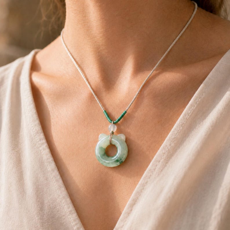 May include: A jade pendant necklace with a circular, cat-shaped pendant. The pendant is light green with darker green marbling. The necklace has a silver chain and green and white accents. The necklace is worn on a person.
