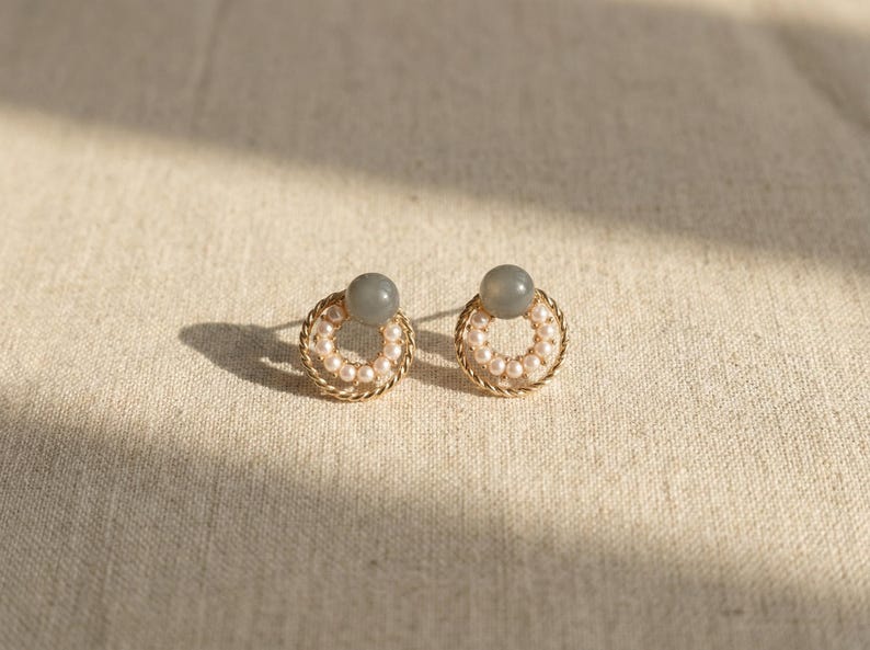 May include: A pair of stud earrings featuring a circular gold-toned twisted metal frame. Each earring is adorned with a ring of small white pearls and topped with a single smooth, round, grey stone bead centered at the top of the circle.