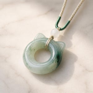 May include: A jade pendant necklace featuring a cat-shaped design. The pendant is a light green color with darker green veining, and is suspended from a beige cord with green accents. A white bead is also included.