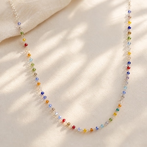 May include: A delicate silver chain necklace featuring a series of small, multi-colored beads spaced evenly along the length. The beads appear in various hues including red, blue, green, yellow, and clear, creating a vibrant, repeating pattern.