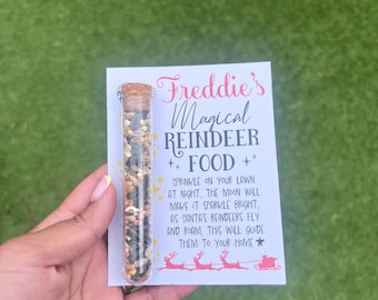Może przedstawiać: Przezroczysta szklana fiolka wypełniona mieszanką nasion i ziaren, zamknięta korkiem, trzymana przed białą kartką. Na karcie napisano "Freddie's Magical Reindeer Food" różowym pismem, z instrukcjami i grafiką reniferów i sań.