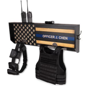 May include: A black wooden wall-mounted shelf with a flag design, featuring a blue stripe and the text "OFFICER J. CHEN." Includes a tactical vest, belt, and two-way radios. A flashlight and hooks are also present.