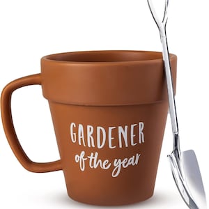 May include: A brown ceramic mug shaped like a flower pot, with the words "GARDENER of the year" in white script. A small, silver shovel-shaped spoon rests against the mug. The mug has a curved handle.