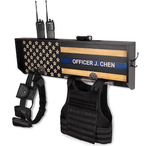 May include: A black wooden wall-mounted gear rack featuring a stylized American flag design with a thin blue line and the text OFFICER J. CHEN. The rack includes metal pipe hardware for hanging a black tactical vest and a black utility belt.