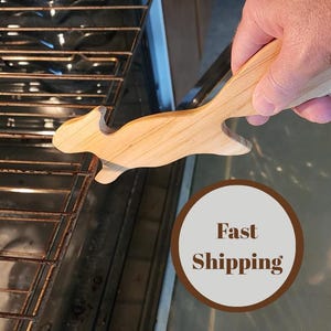 May include: A wooden squirrel-shaped oven rack puller. The puller is light-colored and held by a hand. The oven rack is visible in the background. The text "Fast Shipping" is in a brown circle.