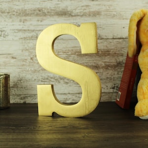 May include: A large, gold-painted wooden letter "S" stands prominently against a light-colored, wood-paneled backdrop. The letter is thick and has a slightly textured surface. A book and a stuffed animal are visible in the background.