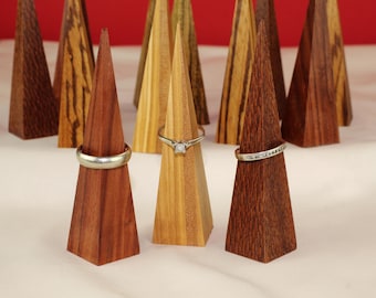 May include: Wooden ring holders in various wood grains, each shaped like a cone. Three holders display silver rings with different designs. The background features additional holders, creating a display of jewelry storage.