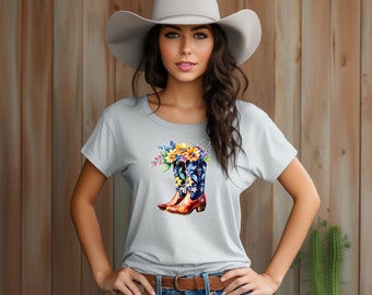 May include: A light gray t-shirt featuring a watercolor design of a cowboy boot filled with colorful flowers. The boots are brown and blue with floral patterns. The model is wearing a cowboy hat and blue jeans.