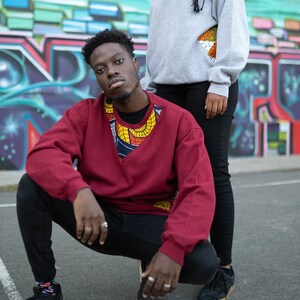 Könnte beinhalten: Zwei Personen posieren vor einer bunten Graffitiwand. Die Person im Vordergrund trägt einen roten Pullover mit einem bunten gemusterten Ausschnitt und eine schwarze Hose. Die Person im Hintergrund trägt einen grauen Pullover mit einem bunten gemusterten Ausschnitt und eine schwarze Hose.