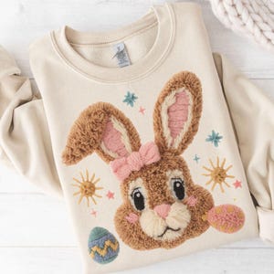 Puede incluir: Sudadera color crema con un diseño bordado de conejo con lazo, orejas y mejillas rosas. El diseño incluye huevos de Pascua y estrellas bordadas. Una prenda festiva y acogedora.