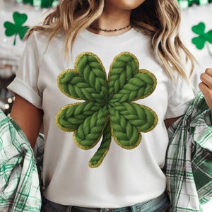 Pode incluir: Camiseta branca com um grande design de trevo de quatro folhas verde e uma borda dourada brilhante. O trevo tem uma aparência texturizada e tricotada. A camiseta faz parte de um tema do Dia de São Patrício.