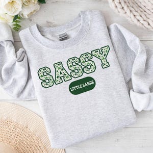 May include: A gray sweatshirt with the word "SASSY" in green letters with a shamrock pattern. The letters are outlined in white. Below the word "SASSY" is a green oval with the text "LITTLE LASSIE" in white.