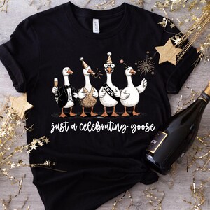 May include: Black t-shirt featuring four cartoon geese dressed for a celebration. One goose wears a tuxedo, another a sequined top, and the third wears glasses and a sash that says "2026". The text "just a celebrating goose" is below.