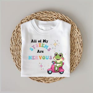 Puede incluir: Camiseta blanca con un gráfico colorido de una rana montando un scooter rosa. El texto "All of My Systems Are Nervous" está impreso en colores arcoíris.