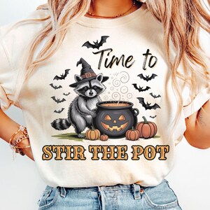 May include: Cream-colored t-shirt with a Halloween-themed graphic. A raccoon wearing a witch hat stirs a cauldron with a jack-o'-lantern face. The text reads "Time to Stir the Pot" with bats and pumpkins.