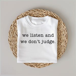 May include: White t-shirt with the text "we listen and we don't judge." printed in black.