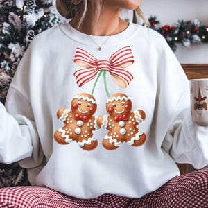 Puede incluir: Sudadera blanca con un diseño festivo de dos hombres de jengibre con pajaritas rojas, conectados a un lazo a rayas. Los hombres de jengibre tienen detalles de glaseado blanco y caras sonrientes, ideal para Navidad.