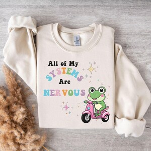 Puede incluir: Una sudadera de color crema con el texto "All of my systems are nervous" en colores arcoíris. Debajo del texto hay una rana de dibujos animados montando un scooter rosa.