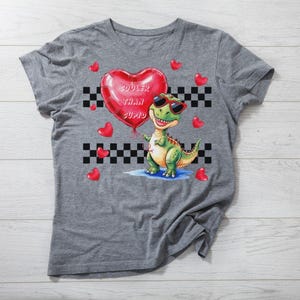 May include: Gray t-shirt with a green dinosaur holding a red heart-shaped balloon that says "Cooler Than Cupid". The dinosaur is wearing sunglasses and there are red hearts scattered around the dinosaur.