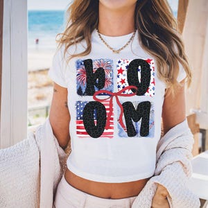 Könnte beinhalten: Weißes, bauchfreies T-Shirt mit einem grafischen Design des Wortes "BOOM" in schwarzen Buchstaben mit einer roten Schleife und einem rot-weiß-blauen amerikanischen Flaggenmuster.