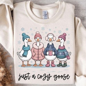 May include: Cream-colored sweatshirt featuring a whimsical illustration of four geese dressed in winter attire. Each goose wears a hat, scarf, or earmuffs, and some have coats and boots. The text "just a cozy goose" is written in a script font.