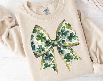 May include: A beige sweatshirt with a green and white shamrock bow on the front. The bow is made of a white ribbon with green shamrocks printed on it.