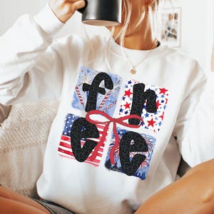 May include: White sweatshirt featuring a graphic design of the word "free" in black letters with a leopard print pattern. The letters are surrounded by red, white and blue stars and fireworks.