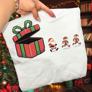 May include: White t-shirt featuring a pixelated red and green gift box with an open lid. Santa Claus and two elves are running to the right. The design has a Christmas theme.