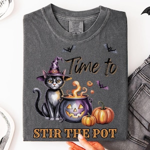 May include: Dark gray t-shirt featuring a black cat wearing a witch hat, sitting next to a cauldron and pumpkins. The text "Time to Stir the Pot" is printed below the image, with bats scattered around the design. The shirt is folded.