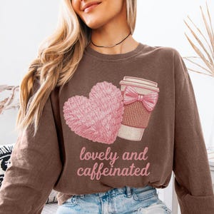 Può includere: Una felpa marrone con un cuore rosa e una grafica di tazza di caffè con un fiocco rosa. Il testo "lovely and caffeinated" è stampato in rosa. La felpa è in stile casual.