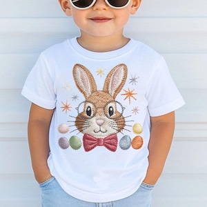 May include: White t-shirt featuring an embroidered Easter bunny wearing glasses and a bow tie. The bunny is surrounded by starbursts and colorful Easter eggs. The shirt is a children's apparel item.