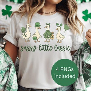 May include: White t-shirt featuring four cartoon ducks dressed in St. Patrick's Day attire, each holding a shamrock or a green accessory. The text "sassy little lassie" is written in green script below the ducks. A green circle with the text "4 PNGs included" is at the bottom.