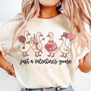 Könnte beinhalten: Cremefarbenes Sweatshirt mit einer Illustration von vier Cartoon-Gänsen, die jeweils mit Accessoires zum Valentinstag geschmückt sind. Die Gänse halten Rosen, einen Pfeil und Bogen, ein herzförmiges Geschenk und einen Ballon. Der Text "just a Valentine's goose" steht darunter.
