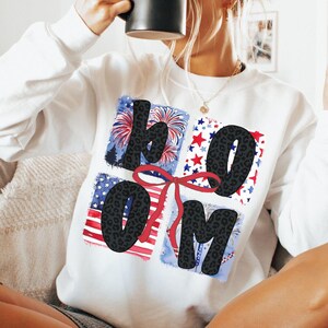 Op de afbeelding: Witte sweatshirt met het woord "BLOOM" in zwarte letters met luipaardprint. De letters staan tegen een achtergrond van rode, witte en blauwe vierkanten met vuurwerk, sterren en strepen. Een rode strik verbindt de "O" en de "O".