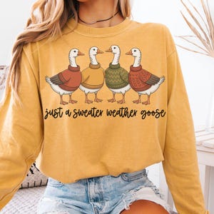May include: Mustard yellow sweatshirt featuring a graphic of four geese wearing sweaters in shades of red, yellow, and green. The text "just a sweater weather goose" is printed below the geese in a black script font. The sweatshirt has long sleeves.