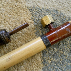 May include: A wooden blowgun with a brass nozzle and a red and black painted section. The blowgun is lying on a brown and gray carpet.