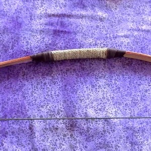 May include: A recurve bow featuring a smooth wooden limb construction with a central grip wrapped in light-colored cordage, flanked by dark brown leather or fabric bindings. A thin, dark string connects the two ends of the bow.