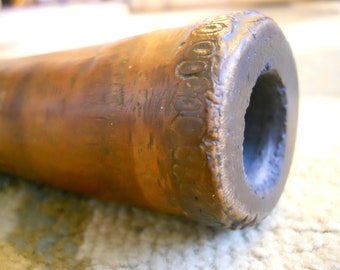 May include: Close-up of a bronze-colored, cylindrical object with a dark opening. The end of the object has a decorative, repeating pattern. The surface shows variations in color and texture, suggesting age or wear. The object appears to be a part of a larger structure.