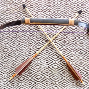May include: A recurve bow with a dark central grip and light-colored limbs, strung with a purple cord, positioned horizontally above two crossed arrows. The arrows feature light wooden shafts, dark brown fletching, and orange-wrapped nocks.