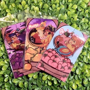 May include: Three colourful acrylic charms in plastic packaging. Each charm features a cartoon character with fruit-themed backgrounds. One charm has the word "Mango" on it. The other charms have images of grapes and peaches.