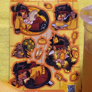 May include: A set of six stickers featuring cartoon monkeys enjoying mango-themed treats. The stickers are yellow, orange, and brown with black outlines. The text "Mango Basket" appears twice on the stickers. A QR code is included on the bottom right corner of the sticker sheet.
