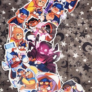Puede incluir: Una colecci&oacute;n de pegatinas de dibujos animados coloridos con personajes parecidos a monos en varias poses. Las pegatinas tienen un borde blanco y est&aacute;n dispuestas sobre un fondo de tela gris con patrones de estrellas y lunas. Los personajes visten trajes en tonos azules, naranjas y amarillos.