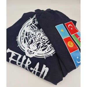 Op de afbeelding: Een donkerblauw t-shirt met een witte wolf-afbeelding en het woord "TURAN" gedrukt op de voorkant. Het shirt heeft ook een rij vlaggen op de rechtermouw, waaronder de vlaggen van Turkije, Kazachstan, Oezbekistan en Azerbeidzjan.