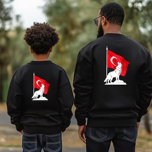 Op de afbeelding: Twee mensen dragen zwarte sweatshirts met een witte wolf die huilt voor een rode Turkse vlag op de rug. De wolf staat op een witte rots.