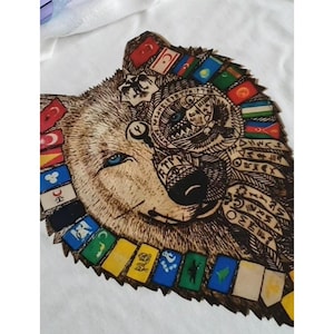Op de afbeelding: Wit T-shirt met een houtgebrand ontwerp van een wolvengezicht. De wolf heeft blauwe ogen en is versierd met vlaggen en ingewikkelde patronen. Het ontwerp combineert natuurlijke houttinten met kleurrijke accenten.