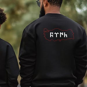 Op de afbeelding: Zwart sweatshirt met een rode omtrek van Turkije en witte tekst erin. De tekst is in een gestileerd lettertype. Het sweatshirt wordt van achteren gezien, met een vage groene boomachtergrond.