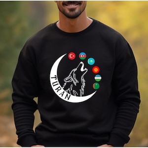 Op de afbeelding: Zwart sweatshirt met een witte wolf die huilt in een halve maan, het woord "TURAN" en verschillende vlagemblemen. Het ontwerp is gecentreerd op de borst.