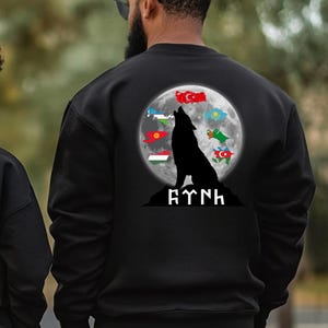 Op de afbeelding: Zwart sweatshirt met een wolf die huilt naar de maan. De maan is omringd door vlaggen van verschillende landen. De tekst "AYTNH" is onder de wolf gedrukt.