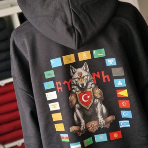 Op de afbeelding: Een zwarte hoodie met een wolf-print op de rug. De wolf draagt ​​een harnas en een rood schild met een halve maan. De print is omringd door kleine vlaggen die verschillende landen vertegenwoordigen. De tekst "RYK" staat boven de wolf en "NH" onder de wolf.