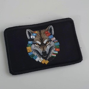 Könnte beinhalten: Ein schwarzer Patch mit einem Wolfskopf in der Mitte. Das Fell des Wolfes ist braun und grau, und seine Augen sind gelb. Der Wolf ist von einem Kreis aus kleinen Flaggen umgeben, die jeweils ein anderes Land repräsentieren. Der Text "RTMA" ist auf die Schnauze des Wolfes tätowiert.